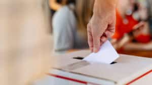 Close-up of hand placing ballot into voting box at election. Ballot and voting box symbolize democracy and elections. Hand placing ballot in election voting box, casting a vote at election