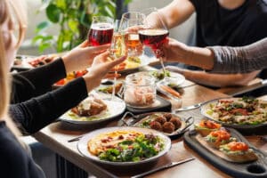 Group of friends clinking wine glasses over a table with gourmet food, celebrating together in a cozy restaurant.